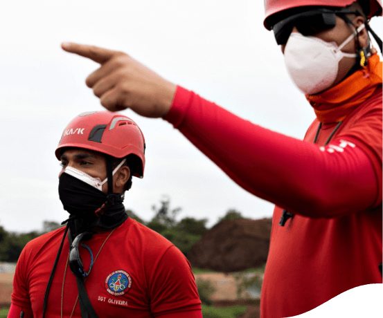 Dois bombeiros estão olhando para o horizonte e, um deles, aponta para frente. Eles estão usando máscara de proteção e capacete.