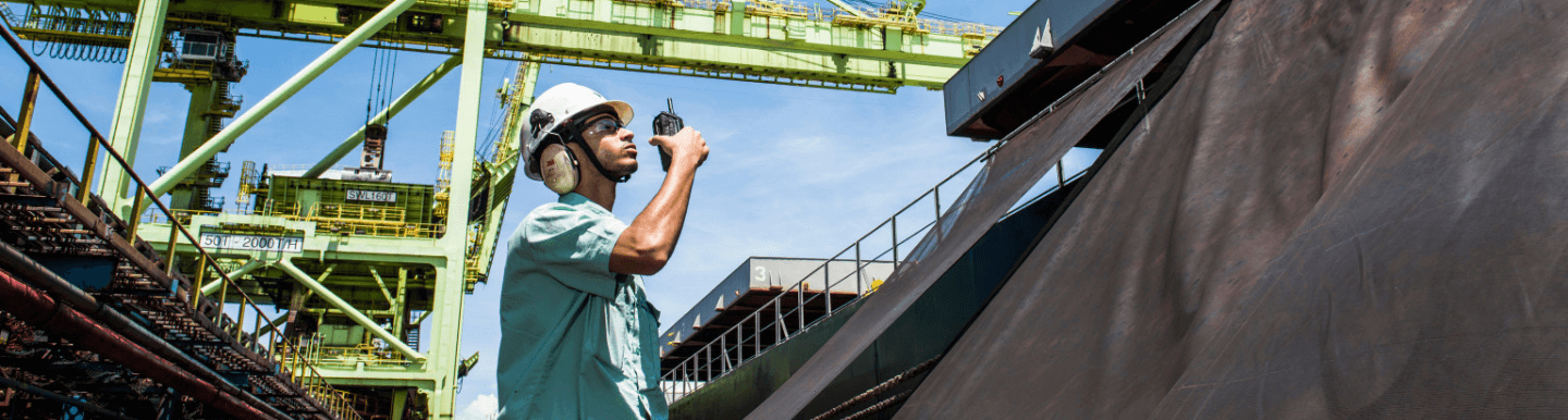 Vale employee in an operational area. He is wearing a light green shirt, goggles, ear muffs and a white helmet, in addition to holding a radio communicator in one hand.