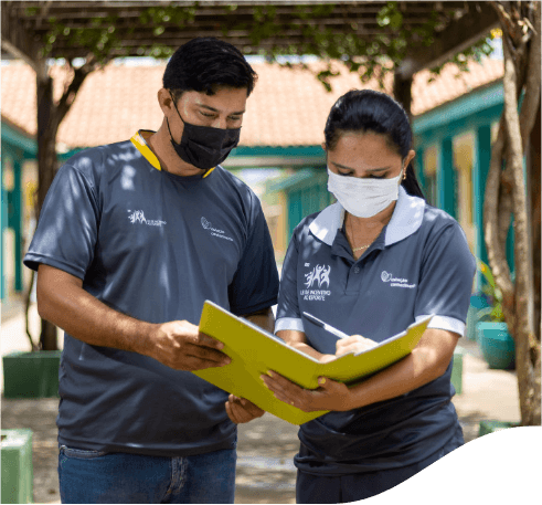 Um homem e uma mulher parados um ao lado do outro escrevendo em um caderno amarelo que ambos seguram. Os dois usam calça jeans, camiseta cinza e máscara facial.