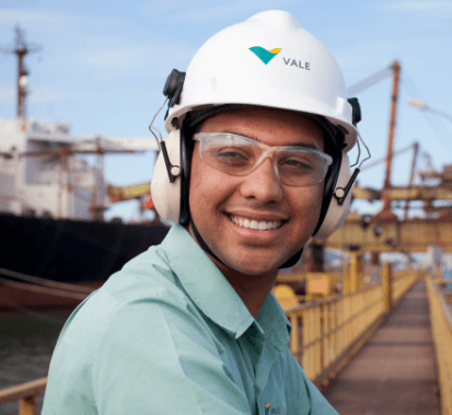 Man smiling in an operations space with a metal structure and a ship in the background. He is wearing a light green shirt, goggles, ear muffs, and a white helmet.