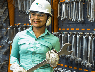 Mulher parada, segurando uma ferramenta de aço em um espaço repleto de outras ferramentas. Ela usa camisa verde clara de uniforme, luvas, óculos de proteção, protetores de ouvido e um capacete branco com logotipo da Vale.