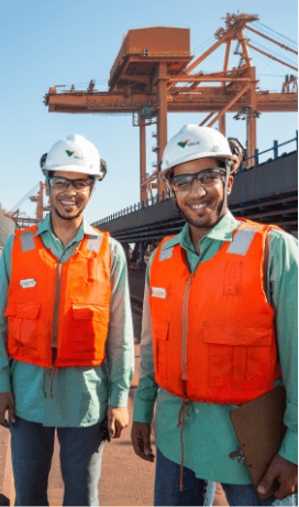 Dois homens sorrindo em uma área operacional. Os dois usam camisas verde de uniforme, colete laranja, óculos de proteção e capacete branco com logotipo da Vale. Um deles segura uma prancheta nas mãos.