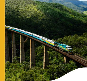 Trem Vale seguindo por uma ferrovia elevada. Em volta é possível ver uma paisagem montanhosa e repleta de vegetação.