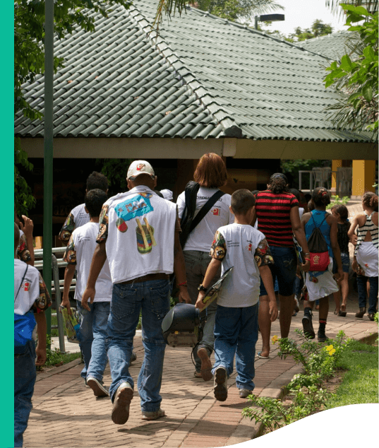 Diversas pessoas estão caminhando, todas de costas para foto. Ao fundo, é possível o telhado de uma estrutura.