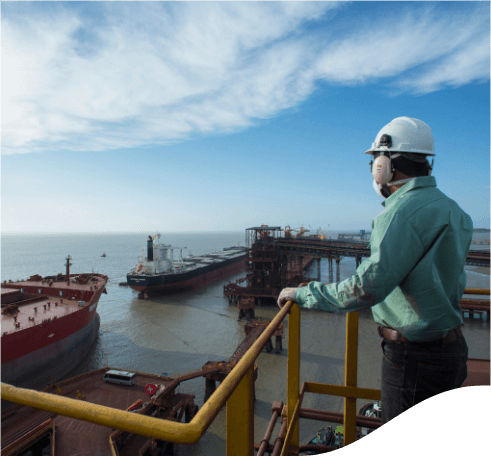 A man on a metal platform, inside a port, looking at a ship in front of him in a lake. He is wearing jeans, a light green shirt, ear muffs, and a white helmet.