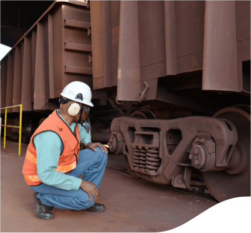 Vale employee crouching beside a large vehicle. He is wearing jeans, a light green shirt, an orange vest, ear muffs, and a white helmet.