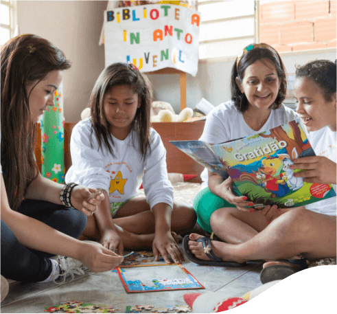 Quatro meninas sentadas no chão lado a lado lendo e pintando revistas infantis. Ao fundo há um cartaz feito com papel cartolina com letras coloridas que diz “Biblioteca Infanto Juvenil”