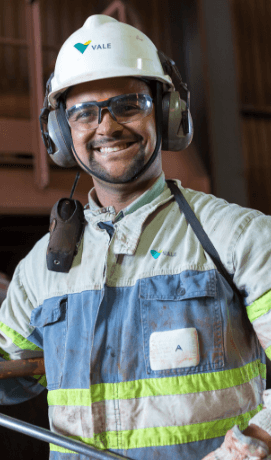 Empregado da Vale sorrindo em uma área operacional. Ele usa uniforme em tons de verde, rádio comunicador pendurado no ombro, óculos de proteção, protetores de ouvido e capacete branco com logotipo da Vale.