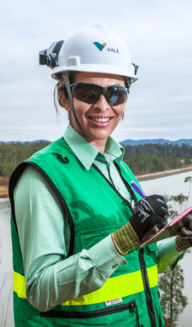 Empregada da Vale sorrindo em uma área operacional. Ele usa camisa verde clara, colete verde, luvas, óculos de proteção e capacete branco com logotipo da empresa. Segura uma prancheta em uma das mãos.