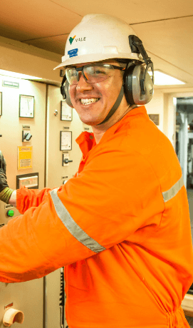 Homem sorrindo em uma área operacional. Ele usa uniforme laranja, óculos de proteção, protetores de ouvido e capacete branco com logotipo da Vale.