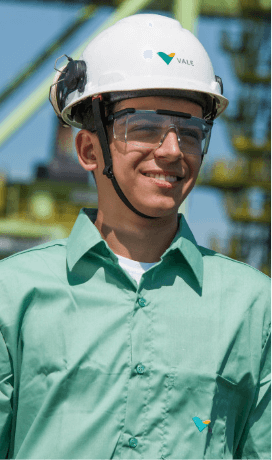 Homem sorrindo em uma área operacional. Ele usa camisa verde clara, óculos de proteção e capacete branco com logotipo da Vale.