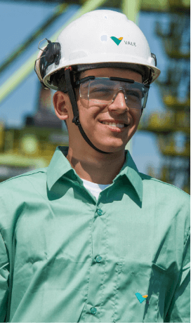 A man smiling in an operational area. He is wearing a light green shirt, goggles and a white helmet with Vale logo.