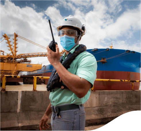 Empregado da Vale parado com um rádio comunicador nas mãos em uma área operacional da empresa ao ar livre. Ele usa calça cinza, camisa verde, máscara facial, óculos de proteção e capacete branco com logotipo da Vale.