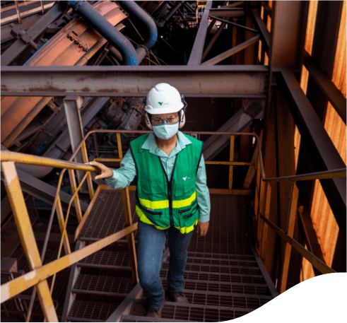 Empregado da Vale subindo as escadas de uma estrutura de metal dentro de uma área operacional. O empregado usa calça jeans, camisa verde clara, colete em um tom mais escuro de verde, máscara facial, óculos de proteção, protetores de ouvido e capacete branco com logotipo da empresa.