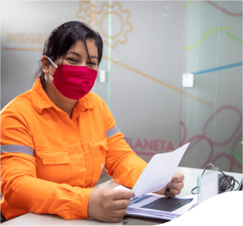 Mulher sentada em uma mesa de escritório. Ela segura uma folha de papel em suas mãos e há outras folhas na mesa. A mulher está usando os cabelos presos para trás, uniforme laranja com detalhes cinzas e máscara facial vermelha.