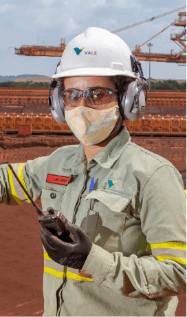 Empregado da Vale em uma área operacional segurando um rádio comunicador em uma das mãos. Ele usa uniforme verde, máscara facial, óculos de proteção, protetores de ouvidos e capacete branco com logotipo da Vale.