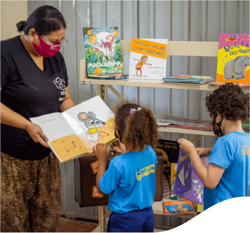 Mulher usando camiseta preta e máscara de proteção facial mostra um livro infantil para uma menina que está a sua frente. A menina está toda vestida de azul e com os cabelos presos. Ao seu lado há um menino também vestido de azul. Atrás deles há uma prateleira com vários livros infantis.