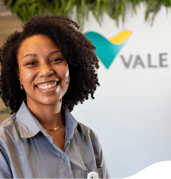 A black woman standing in an office space, wearing a gray shirt and smiling. In the background, there is green and yellow logo of Vale.