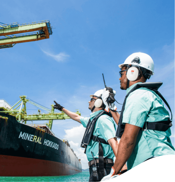 Dois empregados da Vale de frente para um navio em uma área operacional. Os dois usam camisa verde, equipamentos de proteção presos ao corpo, óculos de proteção, protetores de ouvido e capacete branco.