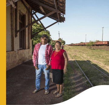 Foto de um casal de idosos sorrindo perto de uma estação de trem. A mulher veste camisa vermelha, saia preta abaixo do joelho e chinelos. O homem está vestindo calça jeans, suéter de tricô branco, camisa xadrez, chinelos e boné.