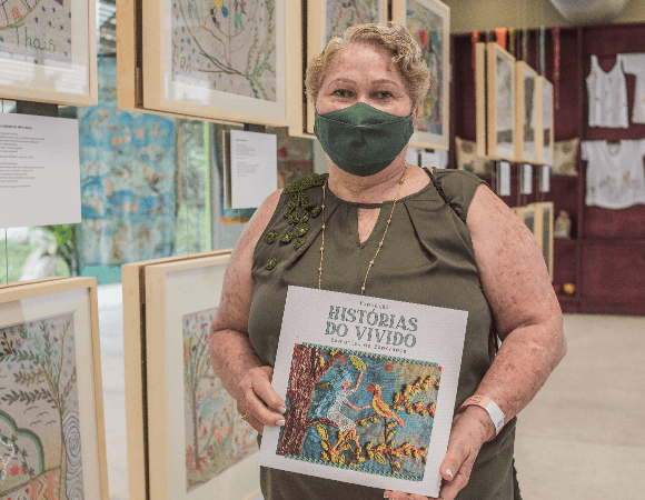 Foto de mulher de cabelos curtos loiros com regata e máscara facial segurando um livro em sua frente. Atrás dela, há vários quadros afixados na parede.