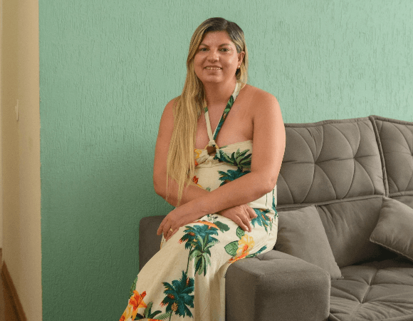 Foto de Flavia Lerman sentada no braço do sofá sorrindo para a câmera. Ela tem cabelos longos, lisos e loiros e usa um vestido longo florido.