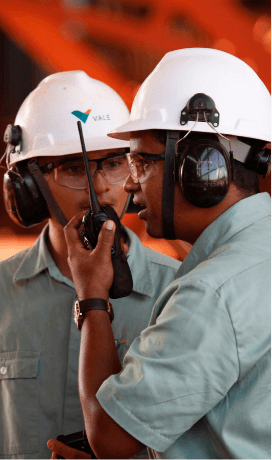 two Vale employees are wearing helmets and goggles. One of them is talking on a radio transmitter.