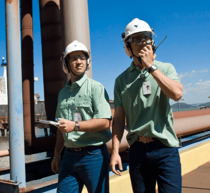 Dois empregados Vale estão em uma área externa de operação. Ambos usam capacete e óculos de proteção e um deles fala através de um radiotransmissor.