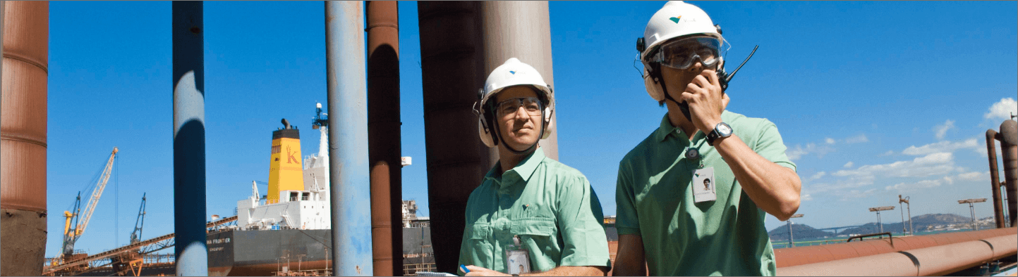 Two Vale employees are in an outdoor operation area. Both are wearing helmets and goggles, and one of them is talking on a radio transmitter.