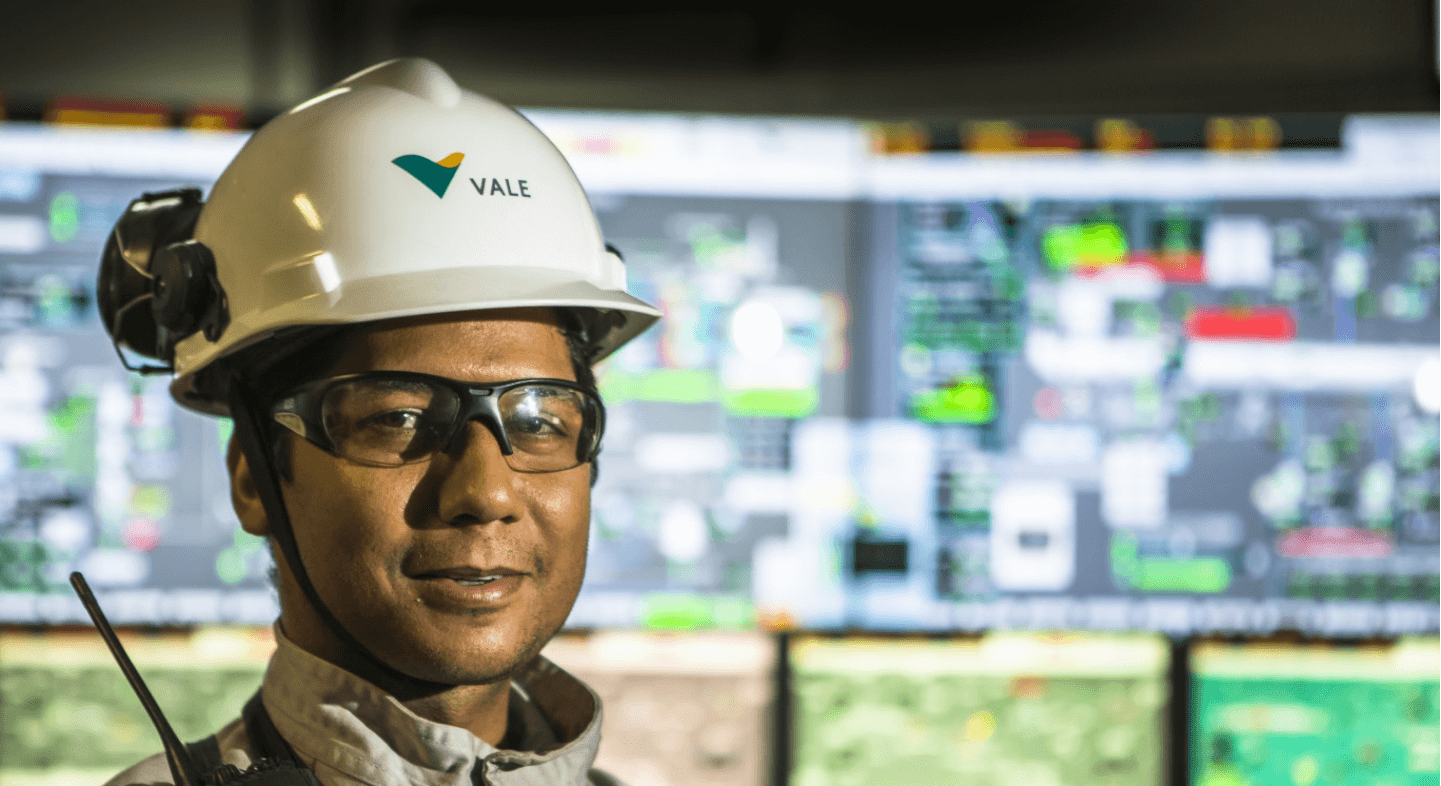 Photo from shoulder height upwards of a Vale employee. He is wearing goggles and a white helmet with the company logo. In the background are several computer screens.