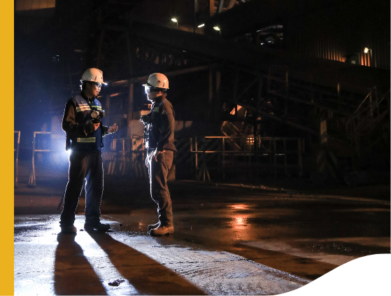 Two men are talking in an outdoor area at night. They are wearing body armor and hard hats.