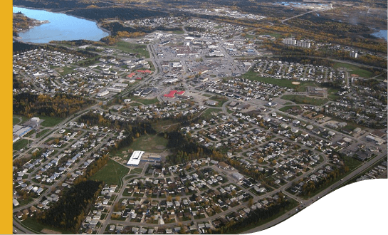 Vista aérea de Thompson. O local tem diversas casas e muito espaço gramado, com exceção do centro, que tem pouca área verde. No canto direito superior da tela é possível ver um lago.