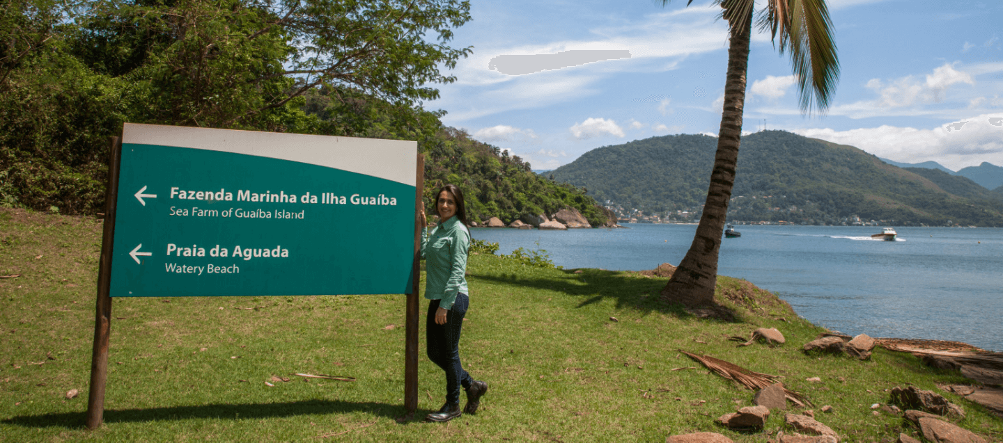 Photo of a woman smiling next to a sign with a coconut tree and one in the background. The woman is wearing Vale uniform, a green long-sleeved button-down shirt, dark pants and sneakers.