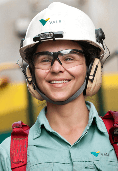 Mulher sorrindo em uma área operacional. Ela usa camisa verde clara com logotipo da Vale, equipamento de segurança vermelho preso aos ombros, óculos de proteção, protetores de ouvido e capacete branco com logotipo da Vale.