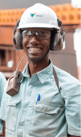 Empregado da Vale sorrindo em uma área operacional. Ele usa camisa verde clara com logotipo da Vale, rádio comunicador pendurado no ombro, óculos de proteção, protetores de ouvido e capacete branco com logotipo da empresa.