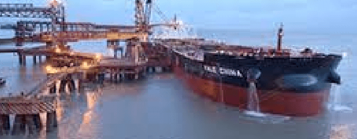 Image of a large cargo ship docked at the port. On the left side, there are several crane-like machines connected to the ship.