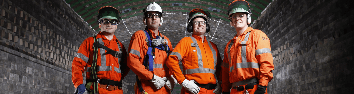 Em um local subterrâneo, quatro empregados Vale – todos de uniformes de proteção laranja, luvas, capacetes e óculos – pousam para foto.