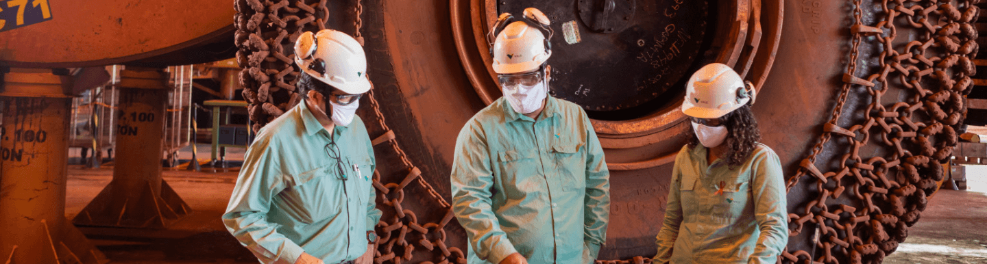 Two men and a woman stand side by side working in an operational space. They are wearing long-sleeved shirts in a light shade of green, helmets, goggles, and protective masks. In the background is a wheel of a large vehicle.