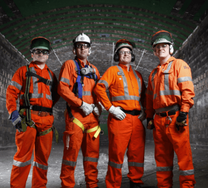 Em um local subterrâneo, quatro empregados Vale – todos de uniformes de proteção laranja, luvas, capacetes e óculos – pousam para foto.