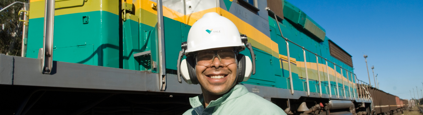 Vale employee smiling. He is wearing a helmet and goggles. In the background is a Vale train, in the colors green and yellow.