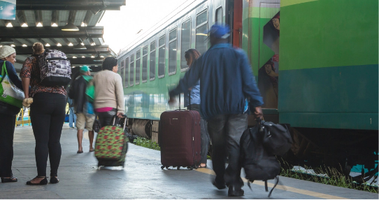 Passageiros com bolsas e malas circulam pela plataforma de trem, enquanto a locomotiva está estacionada.