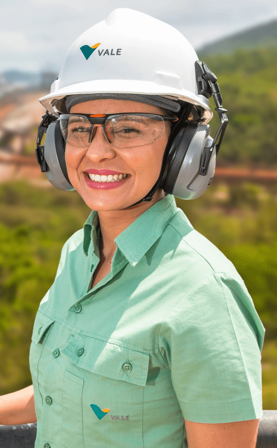Funcionária da Vale sorrindo em paisagem verde. Ela veste uniforme verde
da vale, oculos, capacete e protetores auriculares