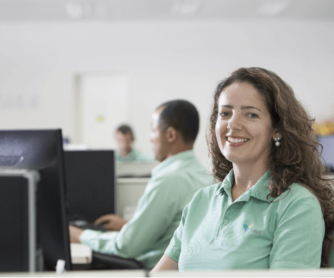 Foto de uma mulher sorrindo em um escritório na frente de um monitor. Ela tem cabelos ondulados, usa brinco de perolas e camisa polo verde clara com o logo da Vale.