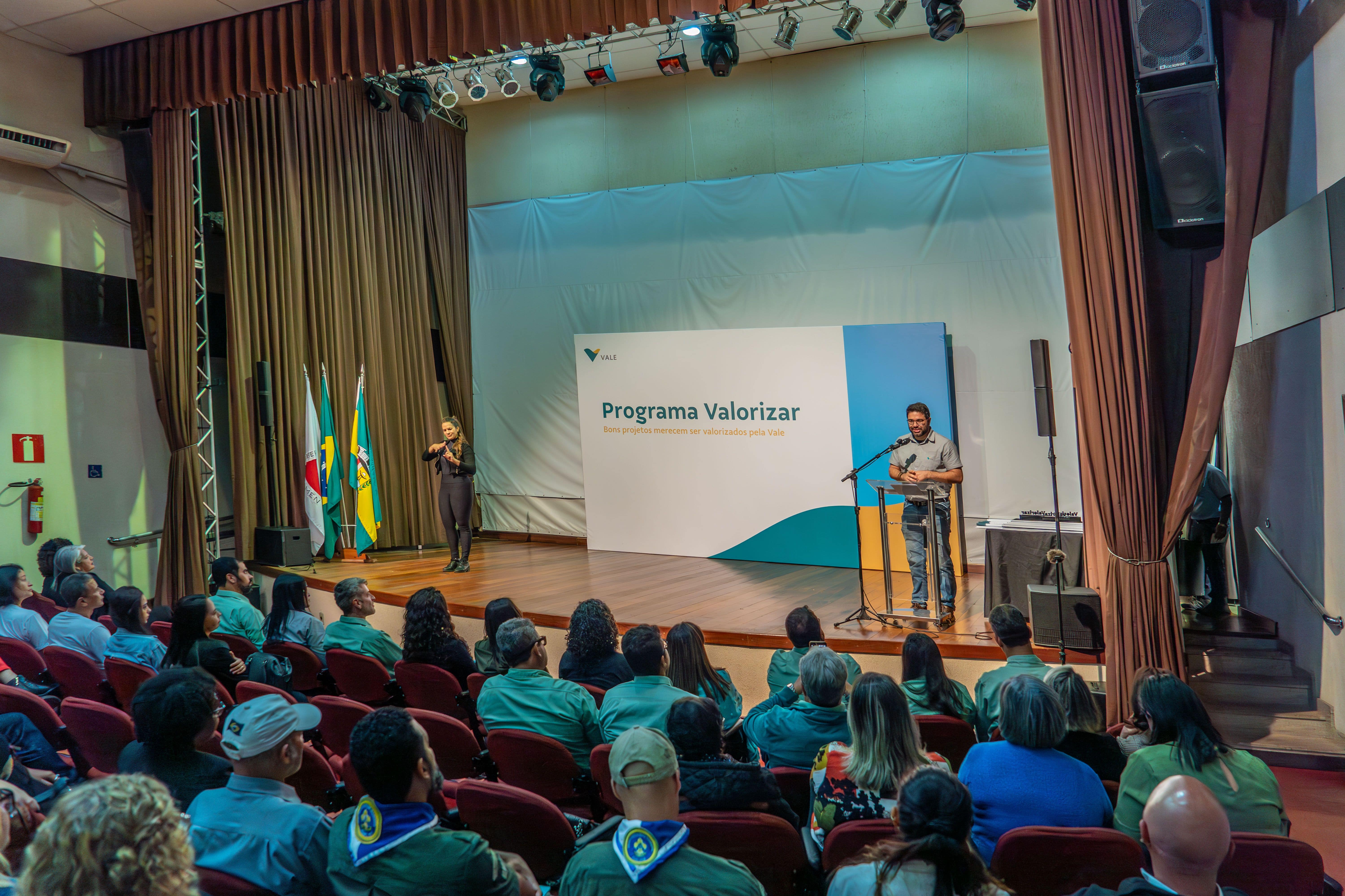 Um empregado Vale discursa em um palco e a plateia está sentada. Há também uma intérprete de libras.