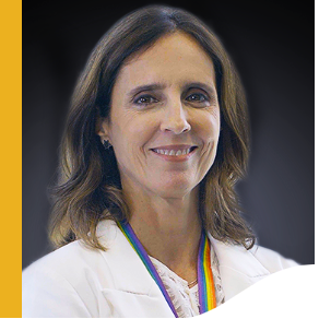 Photo of Marina Quental. She has shoulder-length brown hair, she is smiling and wears a white blazer and a lanyard that holds her badge with the colors of the LGBTQIA+ flag.