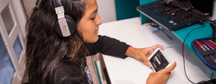 A young woman is sitting, wearing headphones, while using her cell phone.