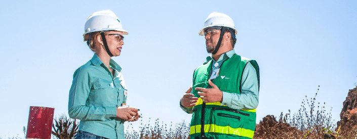 Dois empregados Vale, ambos de capacete de proteção, conversam em uma área aberta, com uma vegetação rasteira ao fundo.