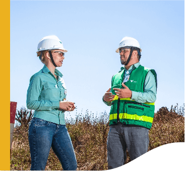 Two Vale employees, both wearing protective helmets, are talking in an open area, with undergrowth in the background.