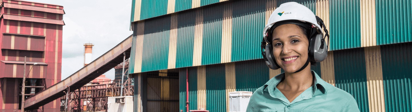 Mulher sorrindo em uma área operacional. Ela usa camisa verde clara, protetores de ouvido e capacete branco com logotipo da Vale.