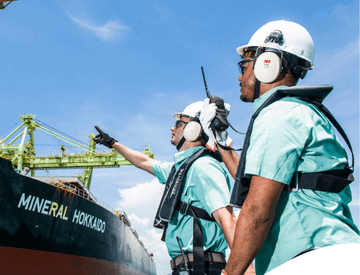 Two Vale employees facing a ship in an operational area. The two are wearing green shirts, protection equipment fixed to the body, goggles, ear muffs, and white helmets.